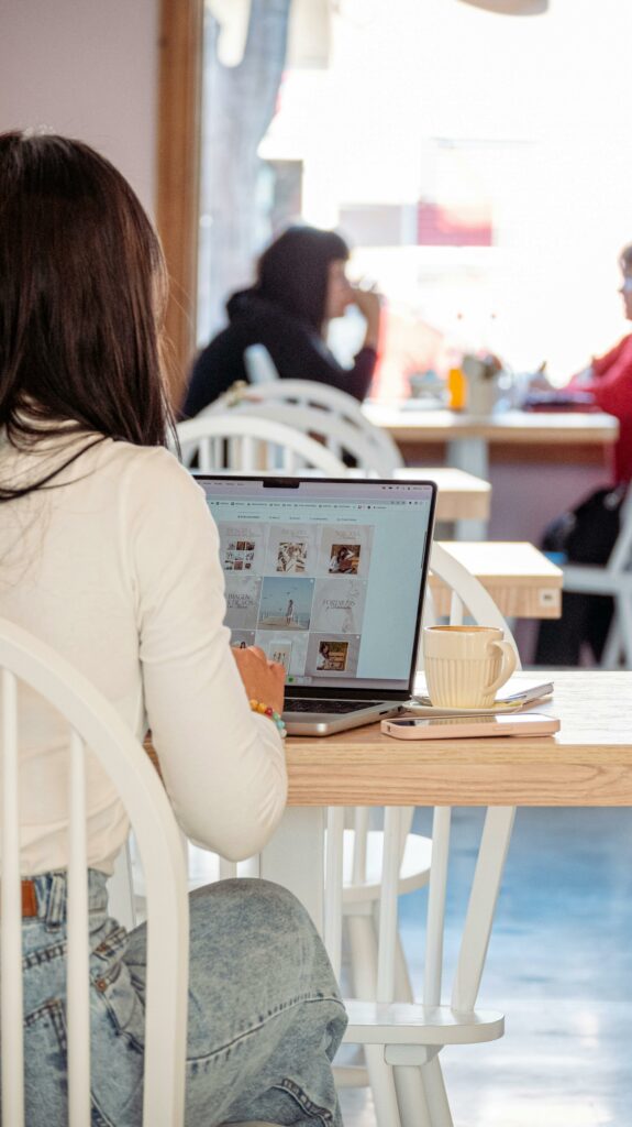 A woman working on a laptop in a cozy café, with a cup of coffee beside her and other people in the background.